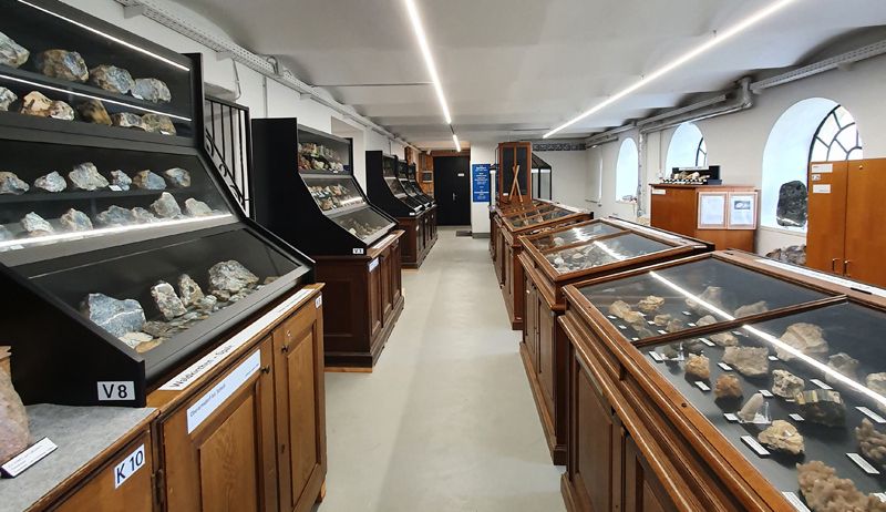 Ein langer Raum mit Reihen von Vitrinen auf beiden Seiten, die verschiedene Steine präsentieren. Jede Vitrine ist mit einer Nummer beschriftet und hat ein weißes Etikett. Die Decke hat Lichter und es gibt bogenförmige Fenster auf der rechten Seite.