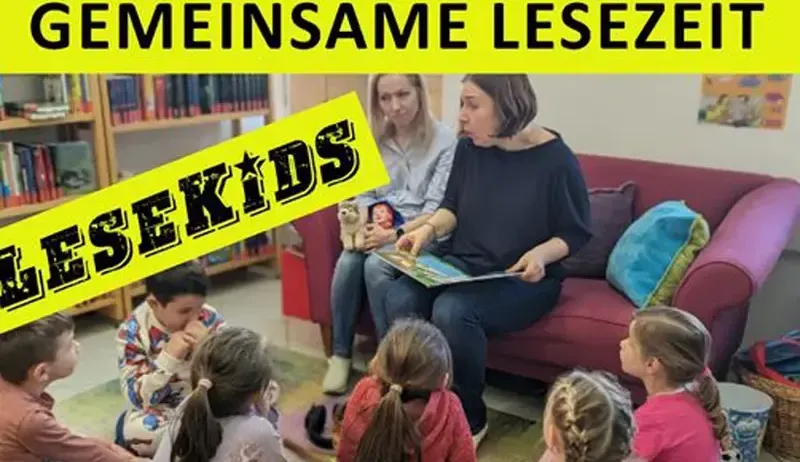 Eine Gruppe von Kindern sitzt auf dem Boden um eine Couch herum und hört einer Frau zu, die ein Buch liest. Die Frau hält ein Buch und schaut die Kinder an. Ein gelbes Schild mit den Worten 'Lesekids' befindet sich links.