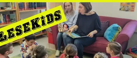 Eine Gruppe von Kindern sitzt auf dem Boden um eine Couch herum und hört einer Frau zu, die ein Buch liest. Die Frau hält ein Buch und schaut die Kinder an. Ein gelbes Schild mit den Worten 'Lesekids' befindet sich links.