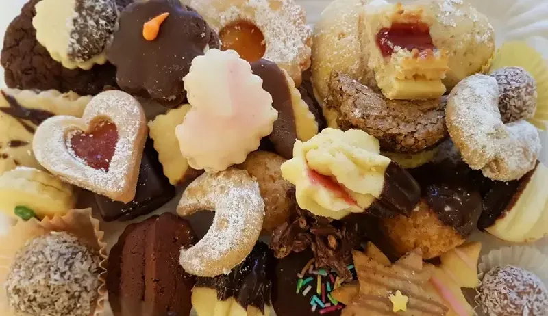 Eine vielfältige Auswahl an Keksen, darunter herzförmige mit roter Füllung, schokoladenüberzogene Sorten und pulverisierte Kekse, ist auf einem weißen Teller angeordnet.