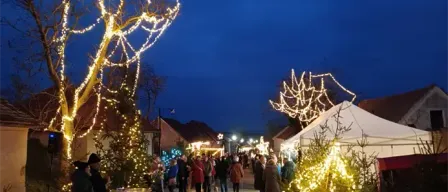 Menschen gehen eine mit Weihnachtsbeleuchtung geschmückte Straße entlang. Rechts steht ein Zelt. Bäume sind mit Lichtern geschmückt, und Gebäude säumen die Straße.