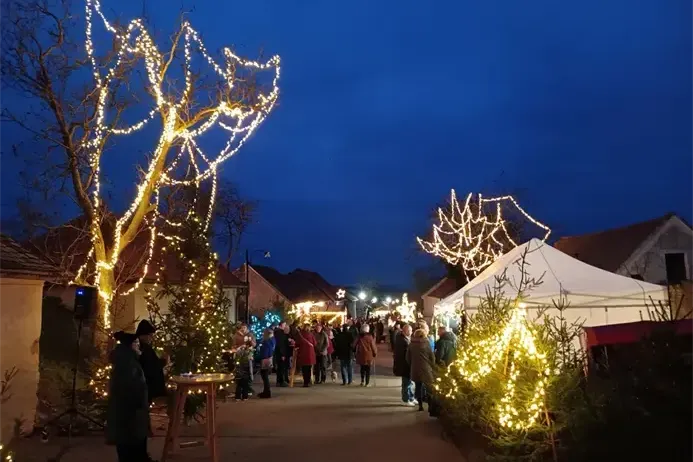 Menschen gehen eine mit Weihnachtsbeleuchtung geschmückte Straße entlang. Rechts steht ein Zelt. Bäume sind mit Lichtern geschmückt, und Gebäude säumen die Straße.
