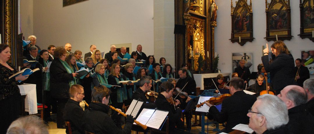 Ein Chor und ein Orchester treten in einer Kirche auf. Der Chor steht vor dem Altar, während das Orchester im Vordergrund sitzt. Sie sind alle formell gekleidet.