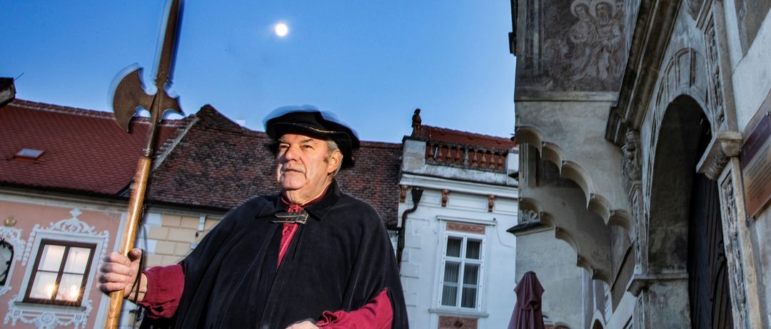 Ein älterer Mann in einem mittelalterlichen Umhang steht auf einem Stadtplatz, hält eine Laterne und eine Hellebarde unter einem blauen Himmel mit Mond.