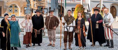 Eine Gruppe von Menschen in mittelalterlicher Kleidung posiert für ein Foto auf einem historischen Stadtplatz mit Gebäuden und einer Statue.