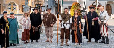 Eine Gruppe von Personen in mittelalterlicher Kleidung steht auf einem Stadtplatz, einige halten Stäbe, und posiert für ein Foto. Dahinter befinden sich Gebäude mit roten Dächern und Statuen.
