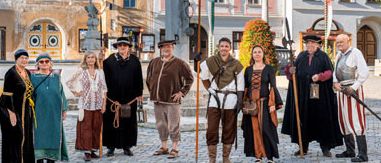 Eine Gruppe von Menschen in mittelalterlichen Kostümen steht auf einem Stadtplatz mit historischen Gebäuden und einer Statue im Hintergrund.