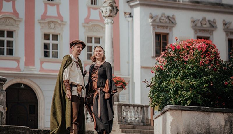 Ein Paar in mittelalterlichen Kostümen steht auf einer Treppe vor einem Gebäude mit kunstvollen Verzierungen und blühenden Blumen.