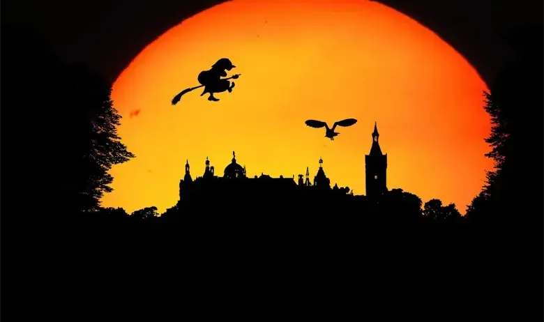 Ein Schattenbild einer Hexe, die auf einem Besen fliegt, und eines Vogels, der gegen einen Sonnenuntergang mit einer Schlosssilhouette im Vordergrund fliegt.