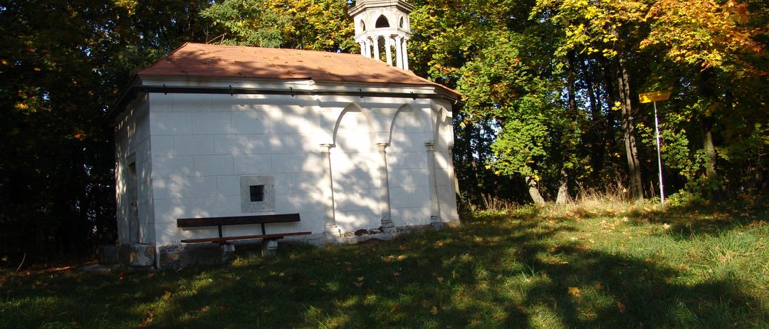 Eine kleine, weiße Kapelle mit einem braunen Dach und Glockenturm steht in einem Wald, umgeben von grünem Gras und gefallenen Blättern.