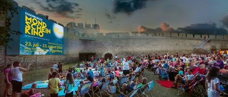 Eine Open-Air-Kinoveranstaltung bei Nacht mit einer großen Werbetafel. Menschen sitzen in Klappstühlen unter einem blauen Himmel mit Wolken.