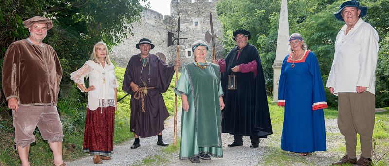 Eine Gruppe von Menschen in mittelalterlicher Kleidung posiert für ein Foto auf einem Kiesweg in der Nähe einer Burg.