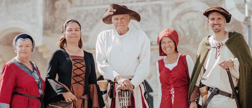 Eine Gruppe von Menschen in historischen Kostümen steht vor einem Gebäude. Sie lächeln und posieren für ein Foto.