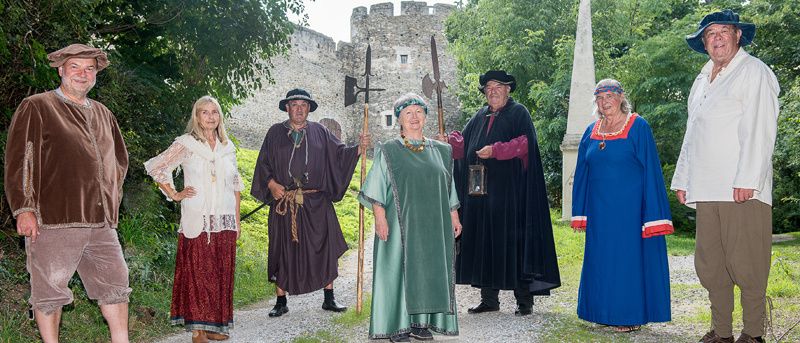 Eine Gruppe von Menschen in mittelalterlichen Kostümen posiert für ein Foto vor einer Burg. Einige halten Speere und eine Laterne.