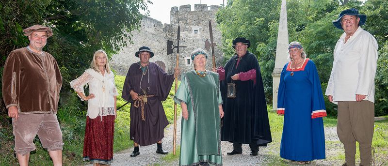 Sieben Erwachsene in mittelalterlicher Kleidung stehen auf einem Kiesweg, drei von ihnen halten Speere. Im Hintergrund befindet sich eine Burg und Grünpflanzen.