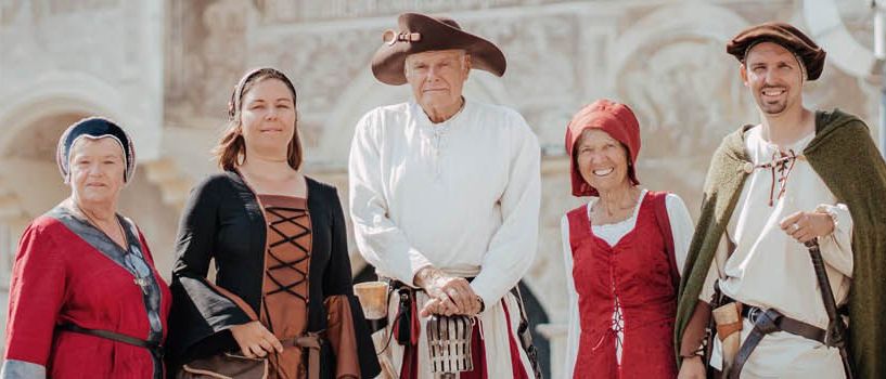 Eine Gruppe von Menschen in historischen Kostümen steht vor einem alten Gebäude. Die Frau in Rot lächelt, und der Mann in der Mitte trägt einen Hut. Die anderen tragen Hüte und tragen verschiedene Gegenstände.
