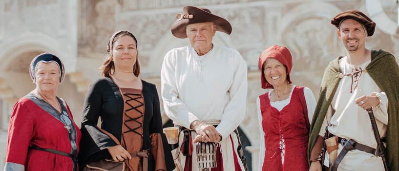 Eine Gruppe von Erwachsenen in mittelalterlichen Kostümen posiert für ein Foto vor einem Gebäude. Sie lächeln und scheinen sich zu amüsieren.