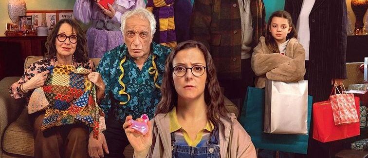 Ein Familienfoto mit Menschen, die Geschenke halten, auf einem weißen Teppich sitzend und stehend. Die Frau in der Mitte hält ein Geschenk mit einer lila Blume. Ein Kind ist vorne, und Taschen und eine Kiste sind auf dem Boden. Das Bild hat ein Filmplakat-Look.