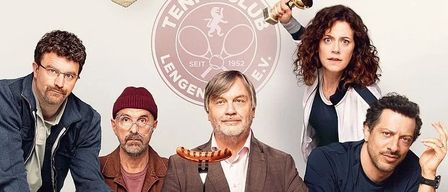 A group of men sit around a table with food. One holds a beer, another a sausage. Behind them, trophies are on a shelf. 'Extrawurst' is written in red.