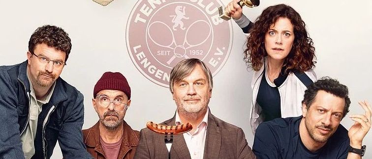 Eine Gruppe von Männern sitzt um einen Tisch mit Essen. Einer hält ein Bier, ein anderer eine Wurst. Hinter ihnen stehen Trophäen auf einem Regal. 'Extrawurst' steht in Rot geschrieben.