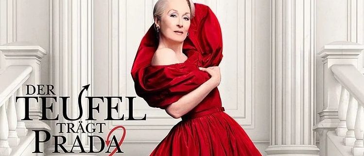 Eine Frau in einem roten Kleid mit Kapuze steht auf weißen Stufen. Der Text im Bild lautet 'Der Teufel trägt Prada'.