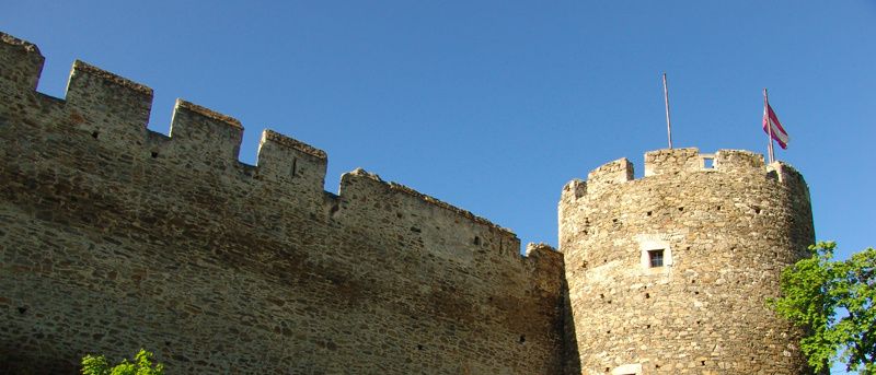 Eine Steinburg mit einer rot-weißen Fahne oben. Der Eingang hat eine Holztür.
