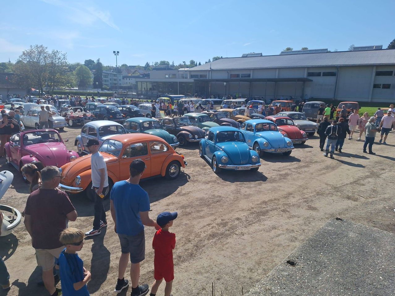Viele Oldtimer sind auf einem offenen Gelände geparkt, auf dem sich Menschen, darunter Männer und Kinder, bewegen. Es gibt auch Straßenlaternen und ein Gebäude im Hintergrund.