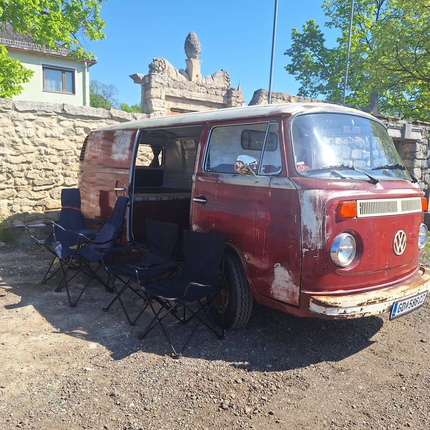 Ein Vintage-VW-Bus in Rot steht vor einer Steinmauer, umringt von mehreren Stühlen. Der Bus hat Rost und Kratzer auf seiner Karosserie.