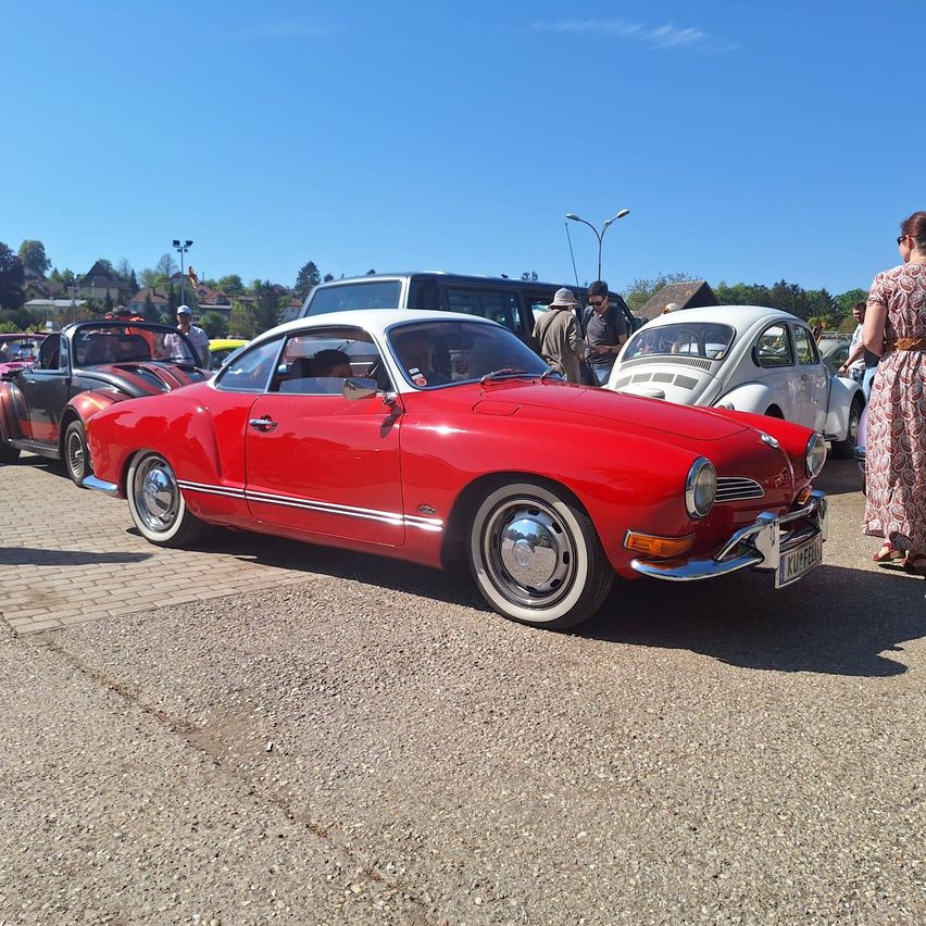 Ein rotes Oldtimer-Auto steht zwischen anderen Oldtimern auf einem Parkplatz. Menschen stehen in der Nähe, einige sind in den Autos.