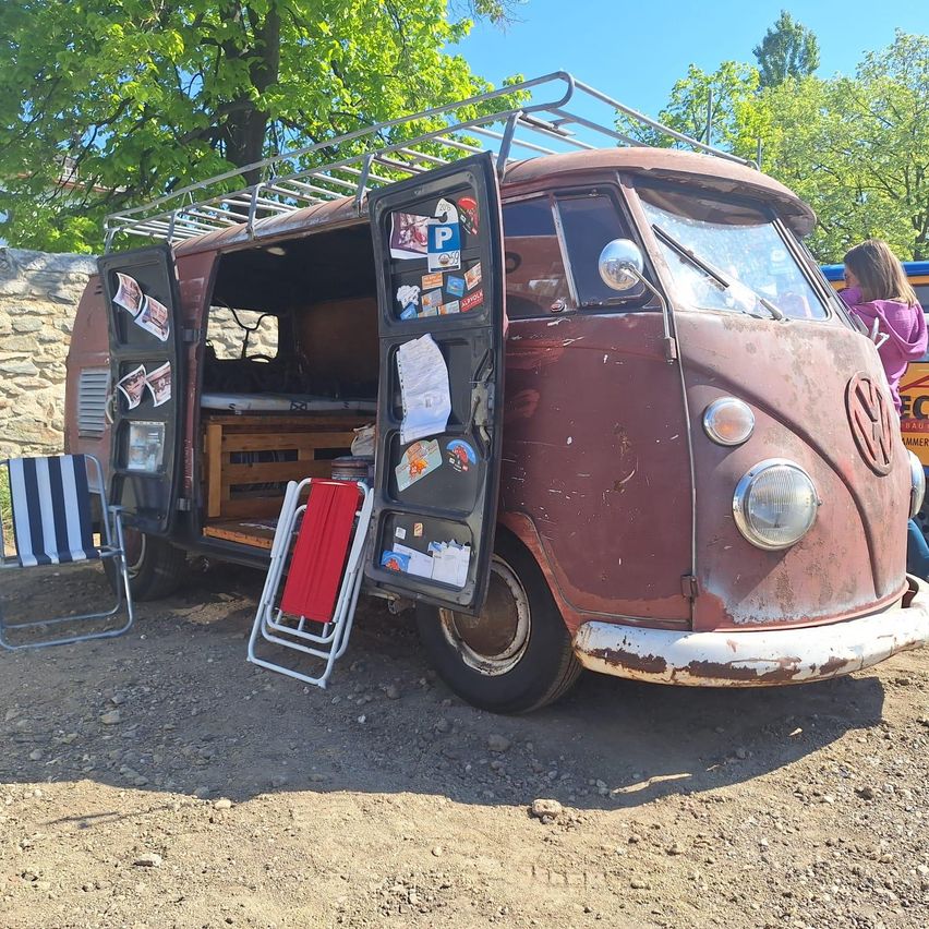Ein alter Volkswagen-Bus ist auf einem Feldweg geparkt, mit offenen Türen. Er hat Aufkleber an den Türen und ein Gepäckträger oben. Ein Stuhl steht neben ihm.