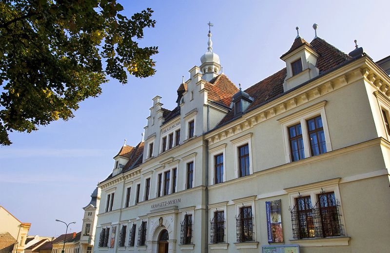 Ein großes, weißes Gebäude mit braunen Dachziegeln und mehreren Fenstern. Ein Baum und eine Straßenlaterne sind im Vordergrund.