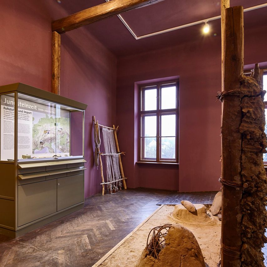 Ein Innenbereich mit einem hölzernen Ausstellungsschrank, einem Informationsschild, einer Holzleiter an der Wand und einem sandigen Bereich mit Steinen.