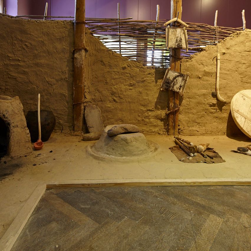 Eine rekonstruierte alte Behausung mit Erdboden, Holzwerkzeugen und einem Lehmofen. Weidenstrukturen und Holzpfähle sind sichtbar.