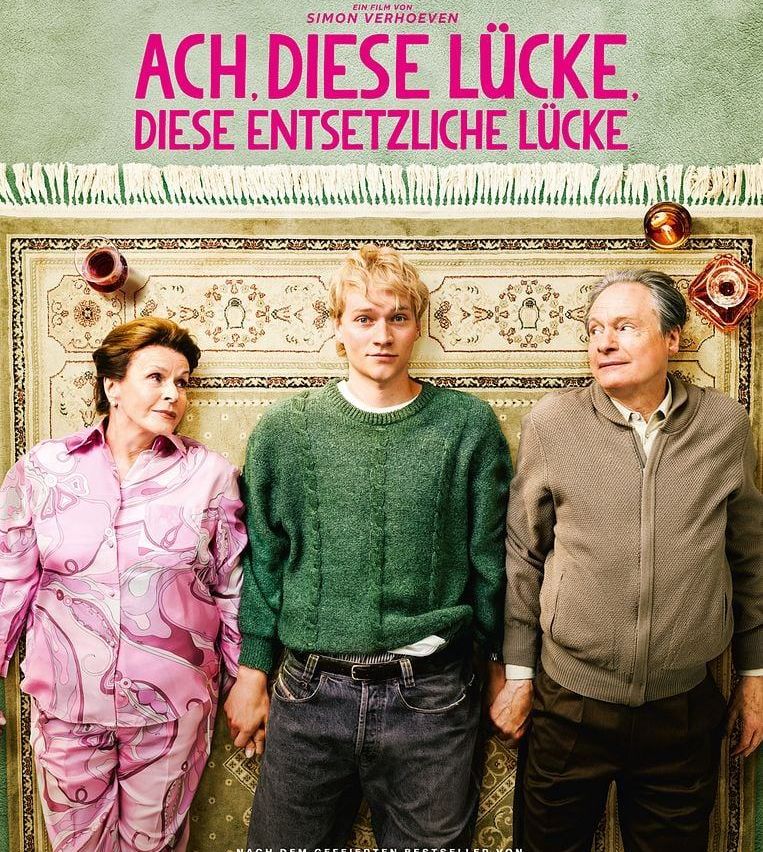 Drei Menschen stehen eng beieinander vor einem gemusterten Hintergrund. Die Frau trägt rosa Pyjamas, der Mann rechts trägt eine braune Jacke und der Mann in der Mitte trägt einen grünen Pullover.