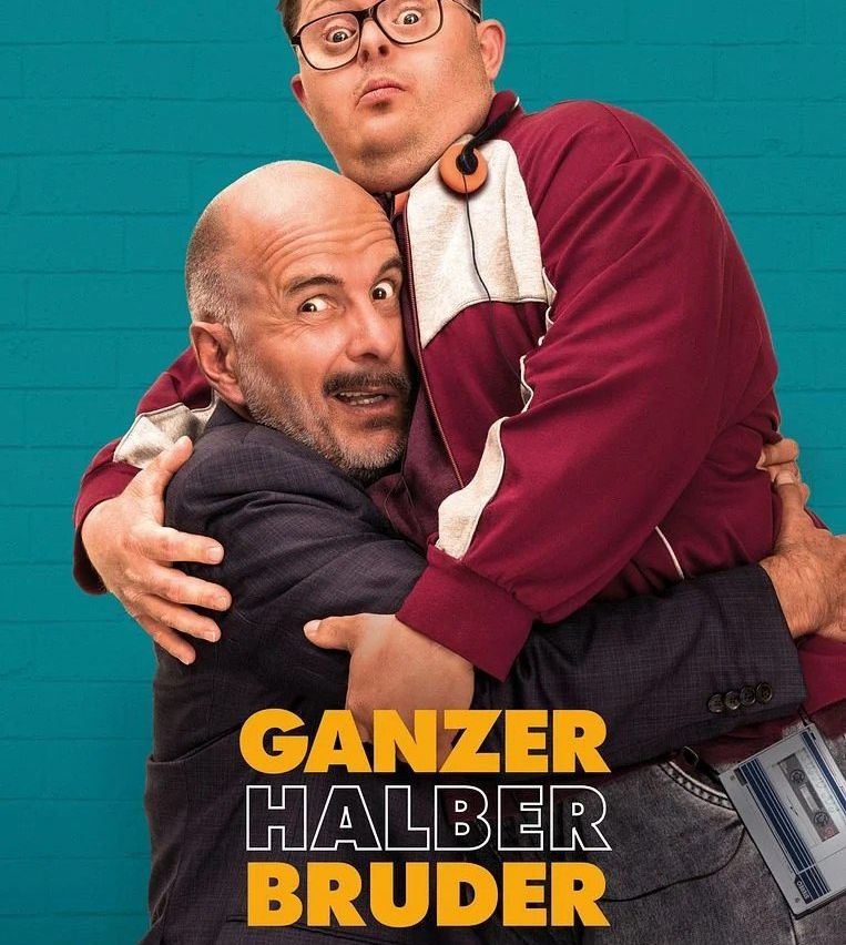 Zwei Männer, einer kahlköpfig und in einem Anzug, der andere in einem burgunderroten Jackett, umarmen sich vor einer türkisen Ziegelwand. Der Text 'GANZER HALBER BRUDER' ist in Gelb angezeigt.