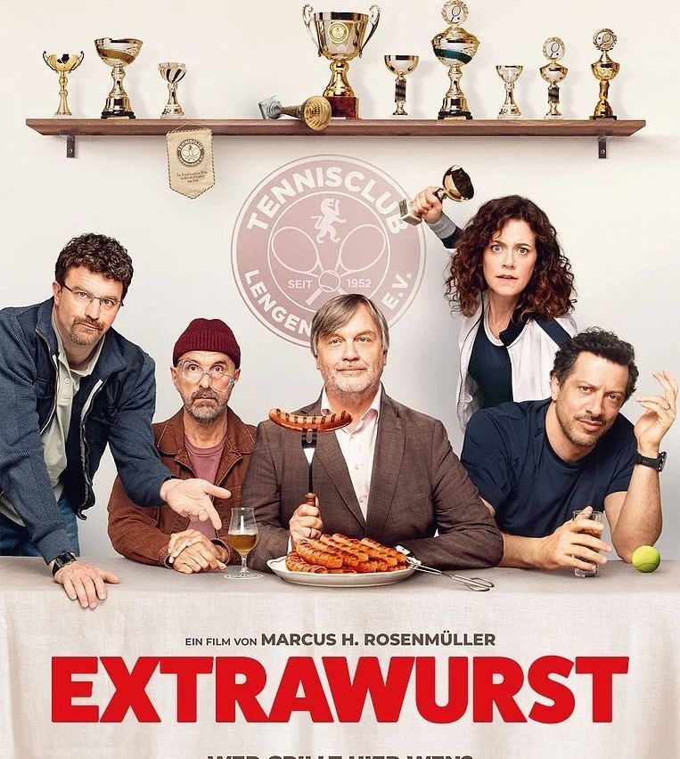 Eine Gruppe von Menschen sitzt an einem Tisch mit Essen und Getränken. Ein Mann hält einen Teller mit Würsten. Hinter ihnen ist eine Wand mit einem Regal mit Trophäen. Der Text 'EXTRAWURST' steht auf dem Tisch.