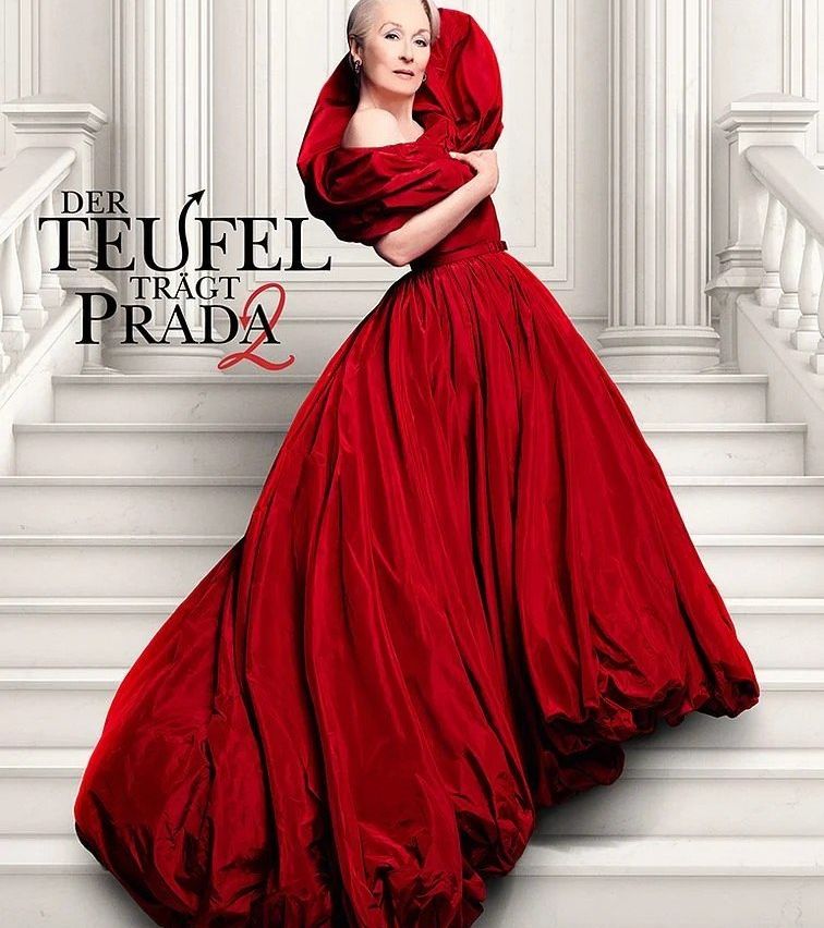 Eine Frau in einem roten Kleid steht auf einer Treppe und hält mit einer Hand den Stoff ihres Kleides. Über ihr steht der Text 'Der Teufel trägt Prada'.