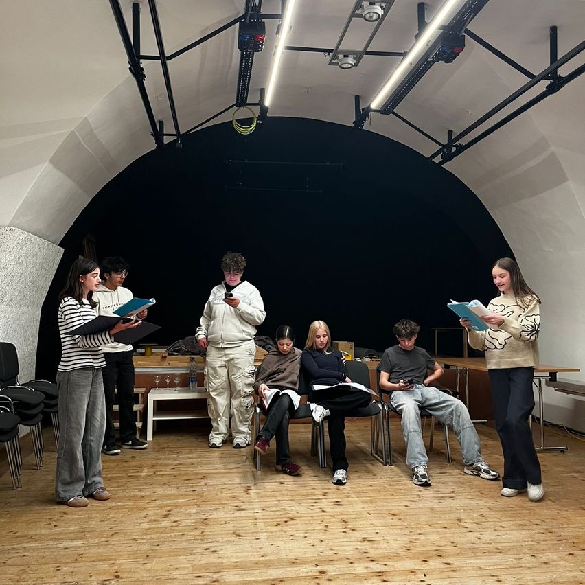 A group of young people are gathered in a semi-circle, possibly rehearsing a play. Some are seated while others stand and hold scripts. The setting appears to be a rehearsal space with a curved ceiling and stage.