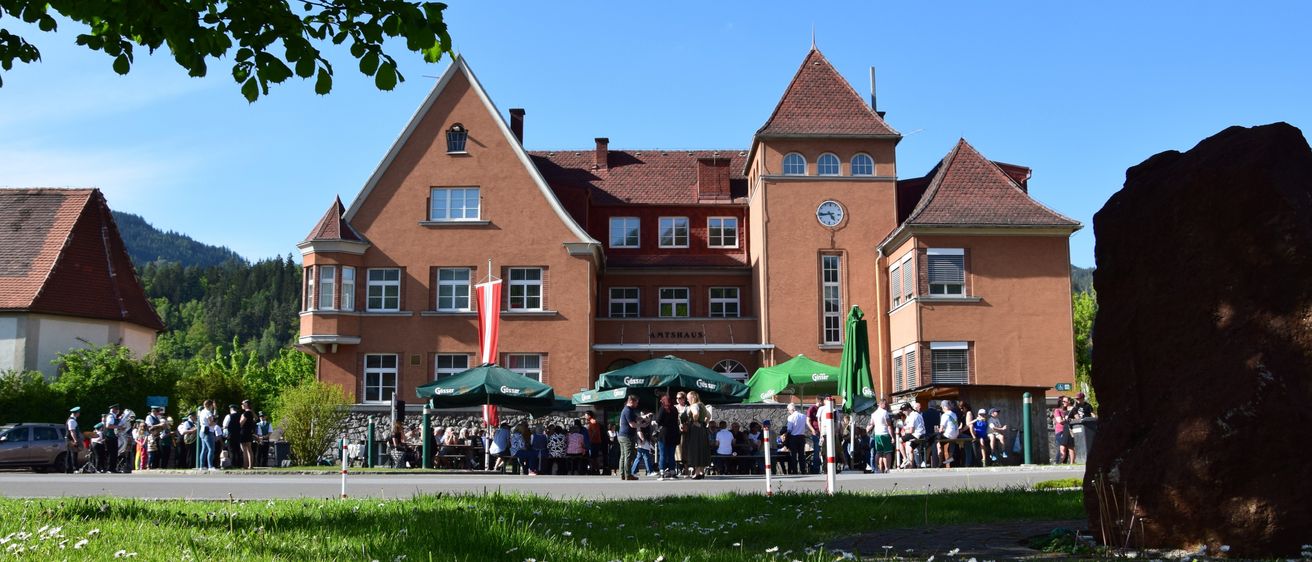Ein großes Gebäude mit braunen Wänden und einem roten Dach steht prominent mit vielen Fenstern. Draußen versammeln sich mehrere Menschen um Tische unter grünen Sonnenschirmen. Die Szene spielt vor einem Hintergrund aus üppigem grünem Gras und Bäumen.