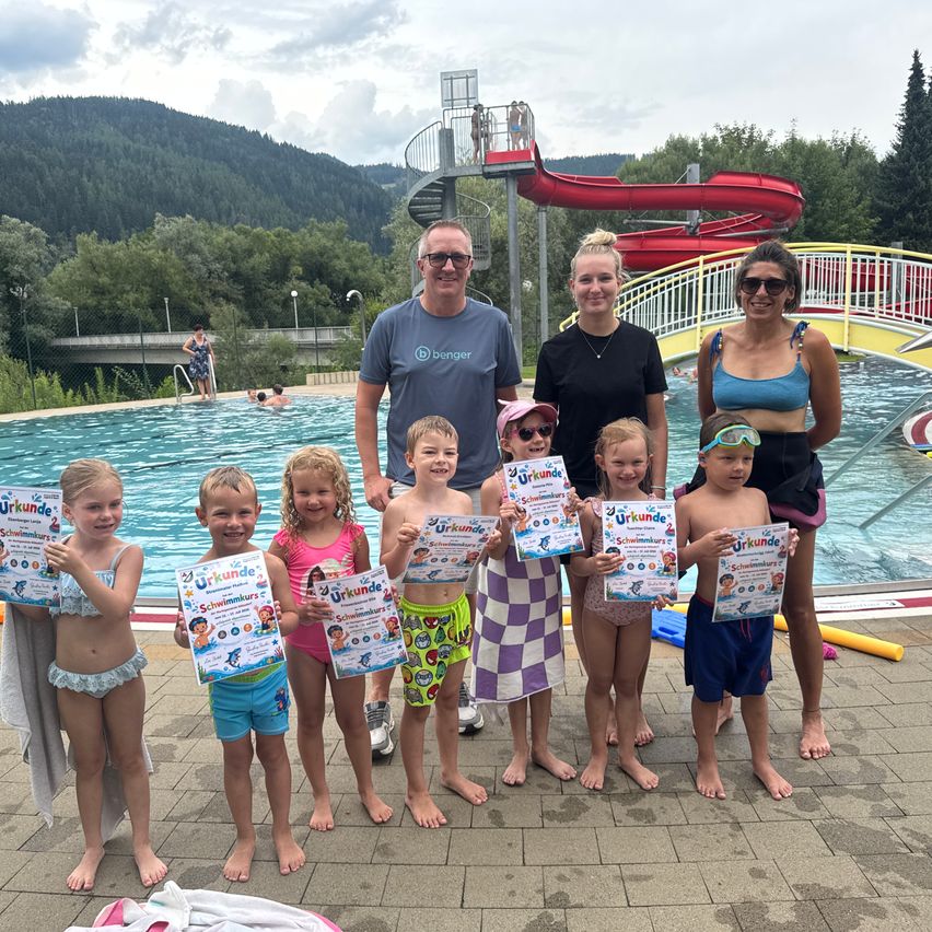 Eine Gruppe von Kindern und Erwachsenen steht vor einem Pool und hält Urkunden in der Hand. Die Erwachsenen lächeln und die Kinder posieren für ein Foto. Dahinter befindet sich ein Pool mit einer Wasserrutsche und einer Brücke. In der Ferne gibt es Berge und Bäume.