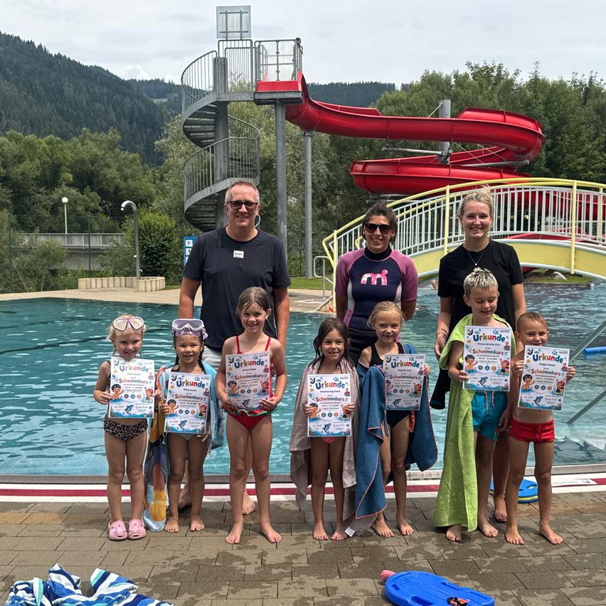Eine Gruppe von Kindern und Erwachsenen steht am Pool, hält Urkunden in den Händen, mit einer roten Wasserrutsche im Hintergrund.