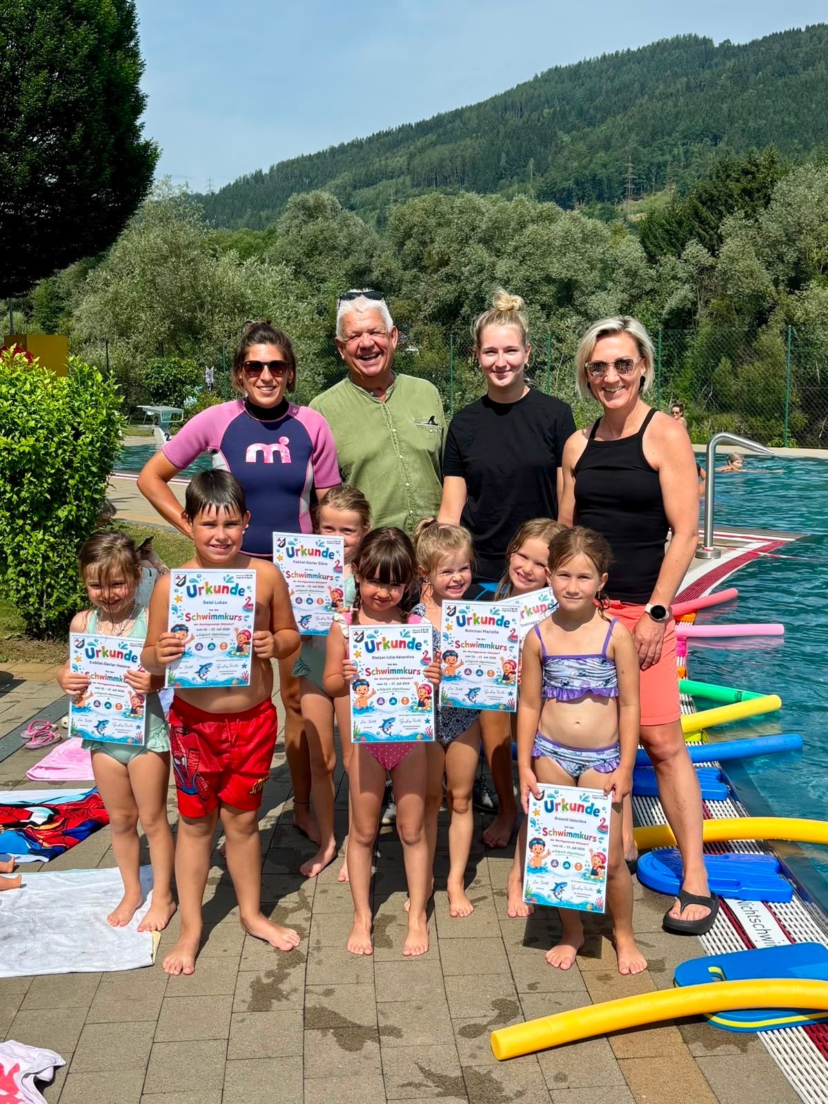 Eine Gruppe von Kindern und Erwachsenen steht am Rand eines Schwimmbeckens und hält Urkunden in der Hand. Die Kinder tragen Badekleidung und die Erwachsenen lässige Kleidung. Das Becken ist von Pflanzen und einem Zaun umgeben. Im Hintergrund befinden sich Berge.