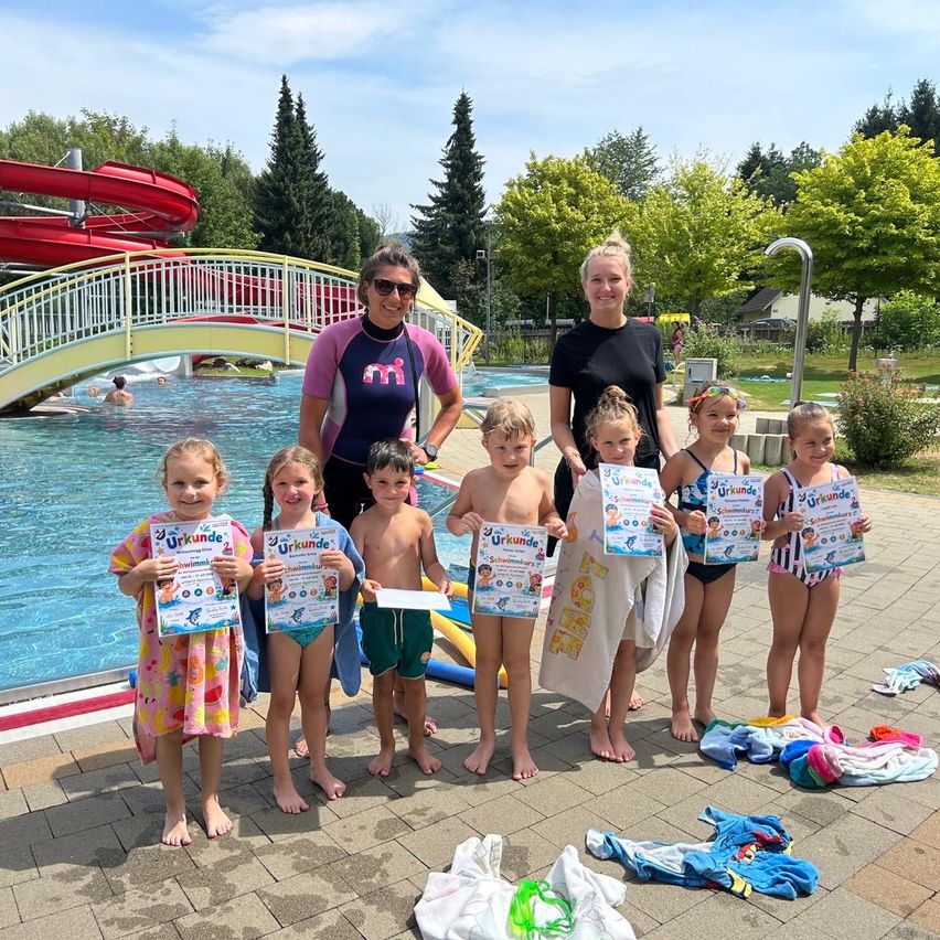 Eine Gruppe von Kindern und zwei Erwachsenen steht am Pool, hält Urkunden und Handtücher. Der Pool hat eine Rutsche und eine Brücke. Bäume und ein Gebäude sind im Hintergrund.