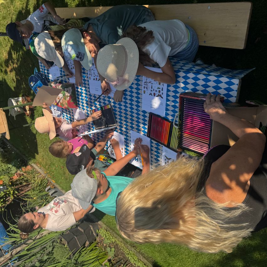A group of children and adults are gathered around a table in a garden, engaged in coloring activities. The table is covered with a blue and white checkered tablecloth, and there are various coloring books and crayons on the table. The children are wearing hats and some are wearing glasses.