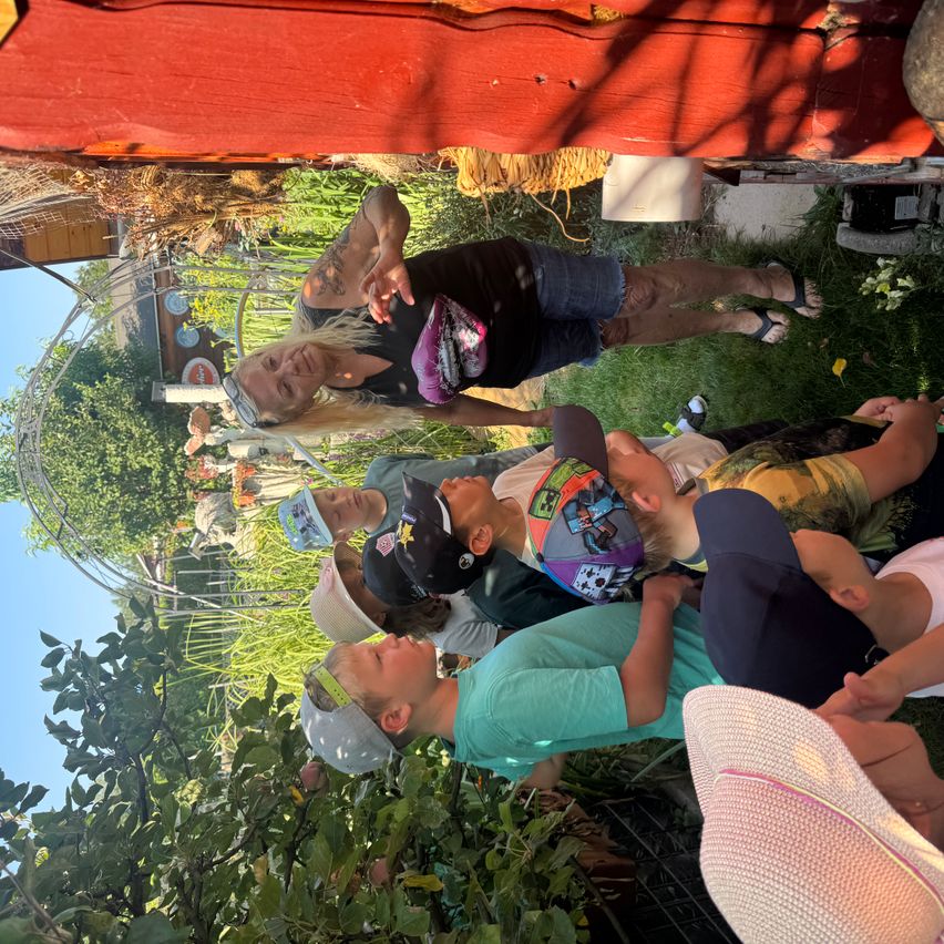A group of people, including children, are gathered in a garden. One woman stands and gestures, while others sit or crouch, some wearing hats. The garden is filled with greenery and a red structure is visible in the background.