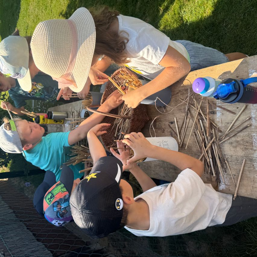 A group of children are gathered around a table, working on a basket-weaving project. One child is using a tool to weave the basket while others assist.