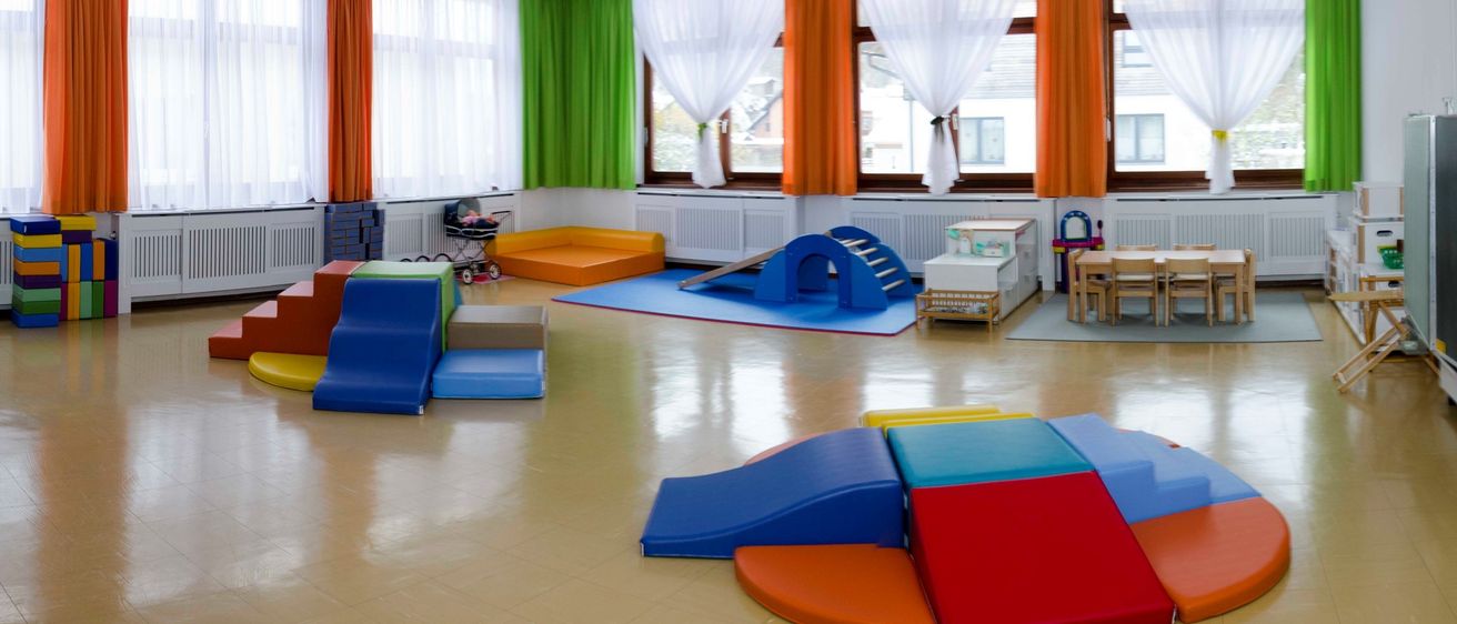 Ein Kinderspielzimmer mit verschiedenen farbenfrohen Spielstrukturen, einem Baby im Kinderwagen und Fenstern mit weißen Vorhängen.