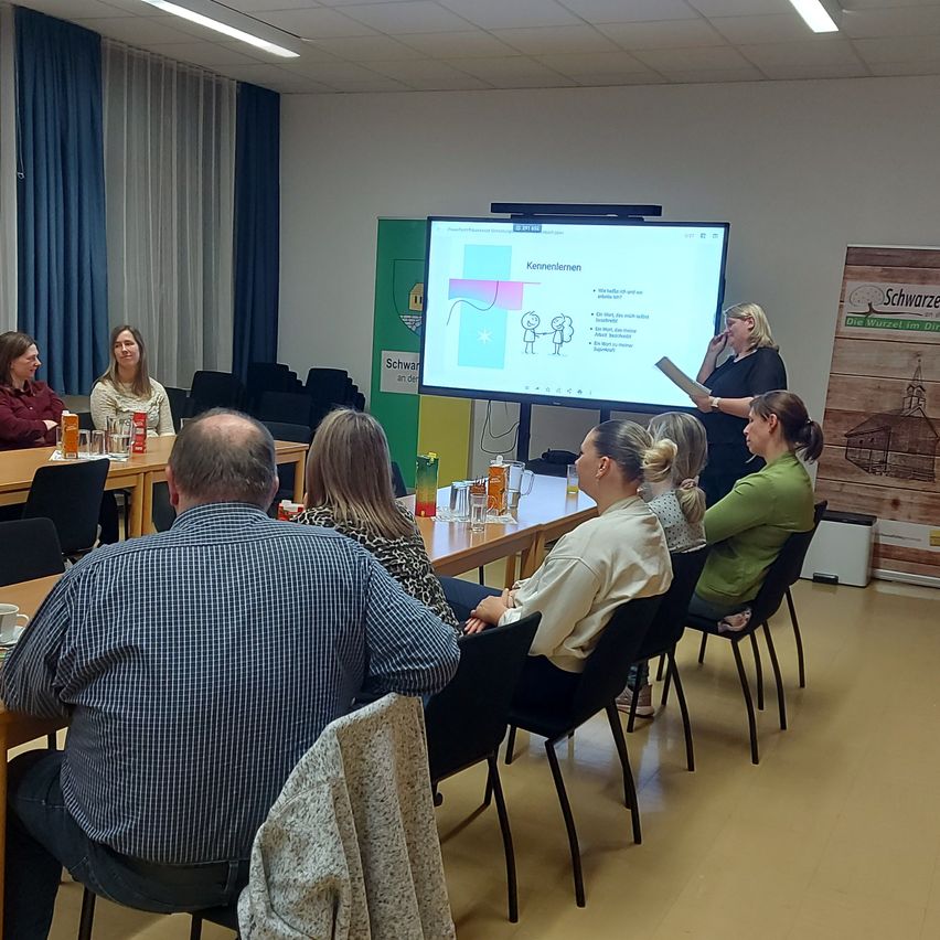 Eine Gruppe von Menschen nimmt in einem Konferenzraum an einer Besprechung teil. Eine Frau steht vor einer Projektionsleinwand, und andere sitzen in Stühlen um einen Tisch mit Tassen und Gläsern.