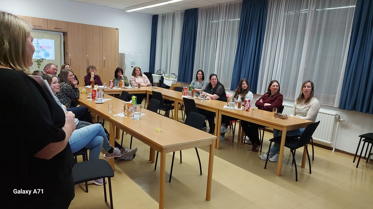 Eine Gruppe von Frauen sitzt in einem Konferenzraum an Tischen. Jeder Tisch hat Getränke und Snacks. Im Hintergrund befinden sich Vorhänge und ein Heizkörper.