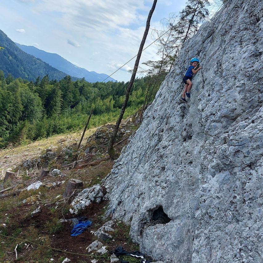 Ein junges Kind in einem blauen Shirt und Helm klettert an einer Felswand mit einem Seil. Das Kind hält den Felsen mit beiden Händen und Füßen fest. Das Gebiet ist von Bäumen und einer Bergkette umgeben.